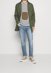 Abrigo verde oliva, camiseta de manga larga a rayas blancas y negras, vaqueros azul claro, bolso cruzado marrón y zapatillas color tostado con suelas blancas.