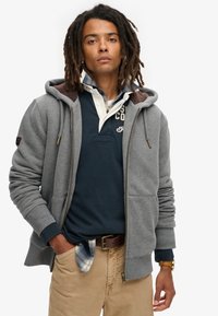 Grijze zip-up hoodie met een fleece-voering in de capuchon, geribbelde manchetten en grote voorkruiszakken, gedragen over een marineblauw polo en beige broek.