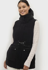 Gilet piumino nero con colletto alto, due tasche frontali, vita con cintura e dettagli in metallo lucido. Indossato sopra una maglia a maniche lunghe con motivo.