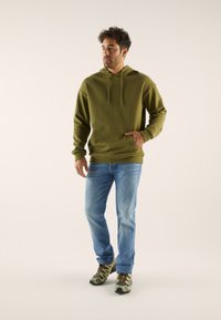 Olive green hoodie with a kangaroo pocket and drawstring hood, paired with light blue denim jeans and camo-patterned shoes.