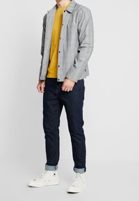 Grå rutig jacka med tryckknappar över en gul sweatshirt, kombinerad med mörkblå jeans och vita högklackade sneakers.