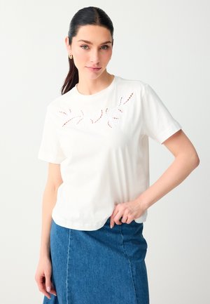 Femme aux cheveux foncés en queue de cheval porte un haut blanc à manches courtes avec des découpes florales en dentelle et une jupe en jean bleue, debout devant un fond blanc.