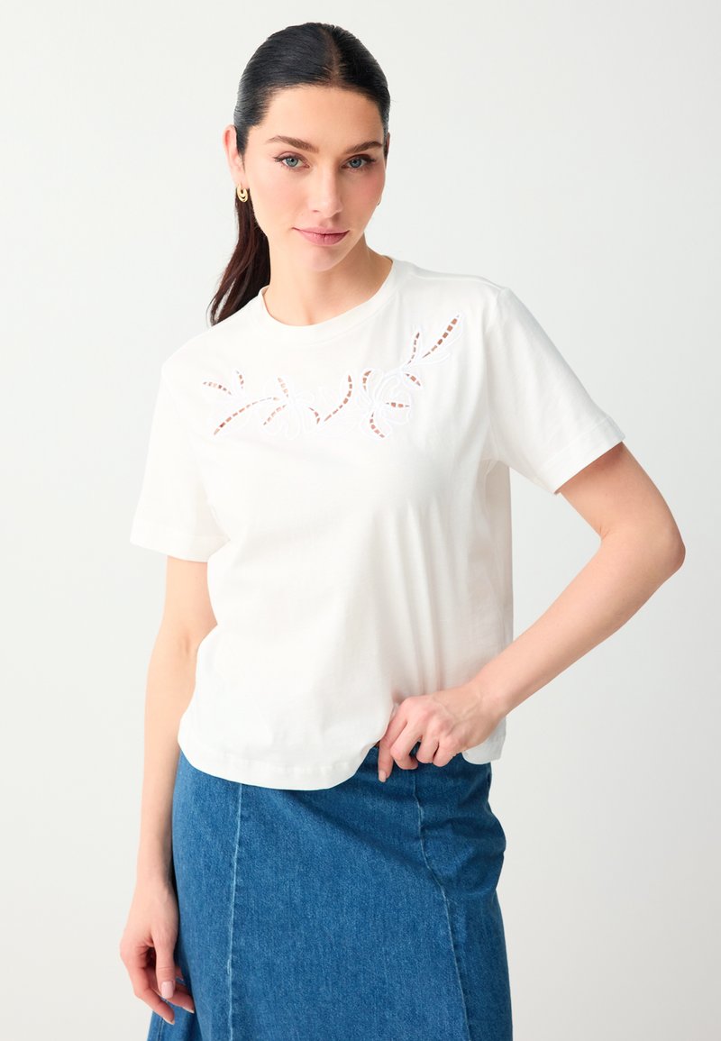 Femme aux cheveux foncés en queue de cheval porte un haut blanc à manches courtes avec des découpes florales en dentelle et une jupe en jean bleue, debout devant un fond blanc.