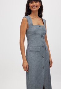 Pilka, prigludusi suknelė su kvadratiniu iškirpimu, trumpomis rankovėmis, priekiniais sagomis ir subtilia tekstūra. Yra dvi kišenės prie juosmens.