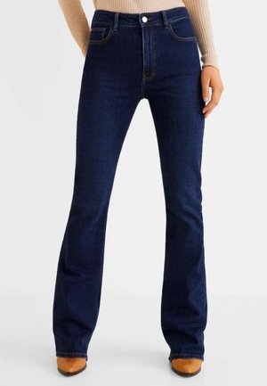 Kobieta ubrana w ciemnoniebieskie dżinsy bootcut, zestawione z beżowym prążkowanym swetrem i brązowymi, szpiczastymi butami, stojąca na jednolitym białym tle.