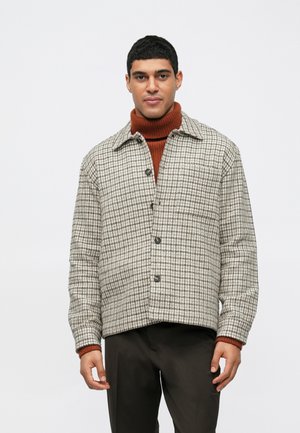 Mann in einem braunen Rollkragenpullover unter einer geknöpften beige-braunen Hahnentrittdame mit dunklen Hosen, der vor einem weißen Hintergrund steht.