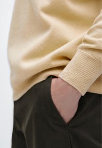 Béžový pletený svetr s žebrovanými manžetami, nošený přes tmavé kalhoty. Ruka spočívá v kapse, čímž se zdůrazňuje hladká texturu látky.