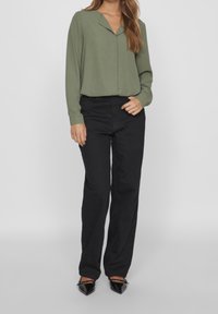 Blusa verde oliva con collo a V, maniche lunghe, abbinata a pantaloni neri a vita alta. Scarpe nere con punta appuntita completano l'outfit.