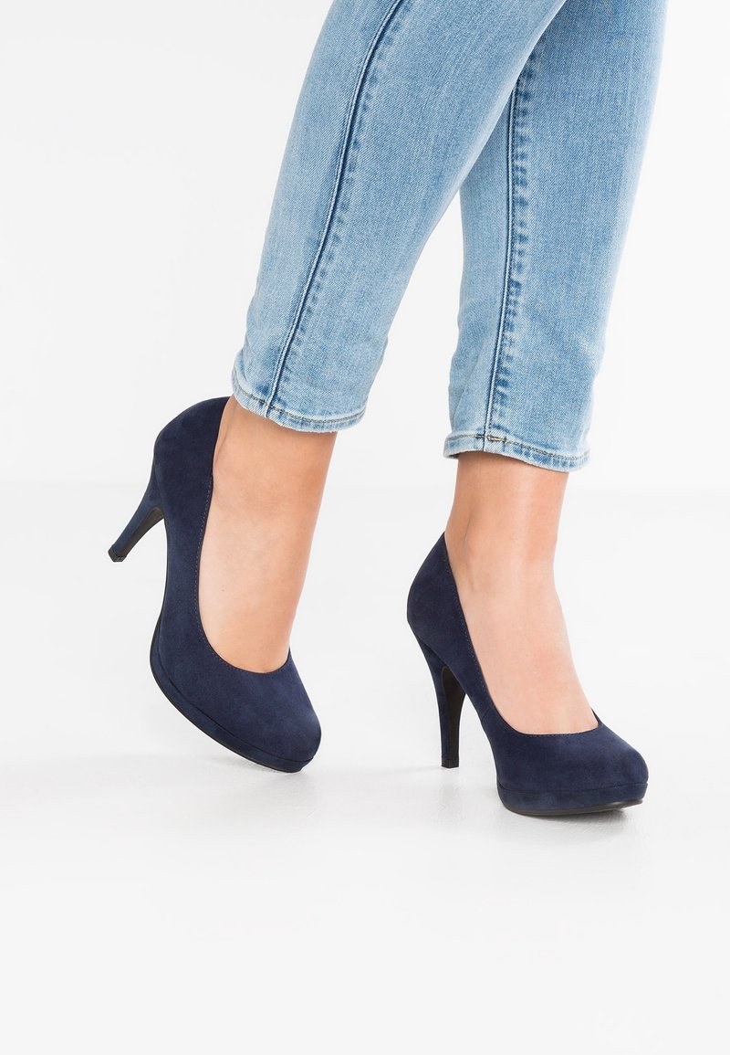 Pieds portant des escarpins en suédine bleu marine associés à un jean skinny bleu clair roulé sur un fond blanc.
