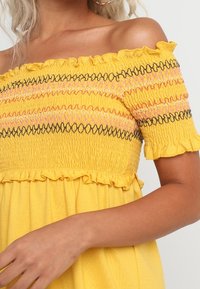 Žluté šaty bez ramínek s volány, nabízející smockovaný živůtek s černým a růžovým cik-cak prošíváním. Měkká, lehká látka.