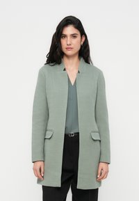 Φωτεινό πράσινο παλτό με υφή και ευθεία γραμμή, που διαθέτει γιακά με σκίσιμο, δύο μπροστινές τσέπες, φορεμένο πάνω από το αντίστοιχο τοπ και μαύρο παντελόνι.