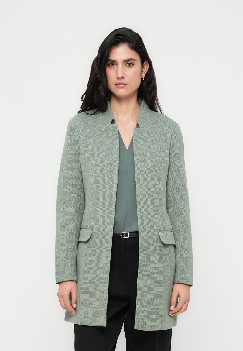 Φωτεινό πράσινο παλτό με υφή και ευθεία γραμμή, που διαθέτει γιακά με σκίσιμο, δύο μπροστινές τσέπες, φορεμένο πάνω από το αντίστοιχο τοπ και μαύρο παντελόνι.