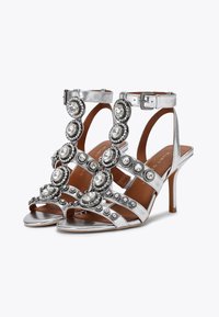 Srebrne, metaliczne sandały na wysokim obcasie z dekoracyjnymi, okrągłymi akcentami z cyrkonii i regulowanym paskiem wokół kostki. Otwarty nosek i smukły design obcasa.