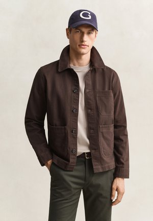Junger Mann trägt eine dunkelbraune, geknöpfte Jacke, beige Hemd, dunkelgrüne Hose und eine marineblaue Kappe mit weißem Buchstaben G.