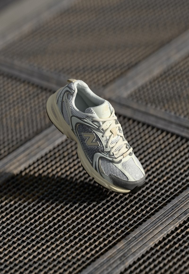 Chaussure de course New Balance blanche et grise avec des détails en mesh reposant sur une surface en grille métallique avec des lignes diagonales.