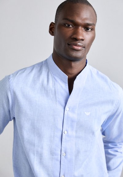 Chemise boutonnée bleu clair avec col mandarin, présentant un motif à carreaux subtil et un petit logo brodé sur la poitrine.