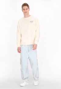 Crème-kleurige sweatshirt met geribbelde bandjes, gecombineerd met lichtblauwe cargobroek met meerdere zijvakken, en witte sneakers.