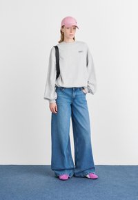 Mujer con gorra rosa, sudadera gris oversized, jeans anchos azules, zapatillas moradas y llevando un bolso negro de hombro, de pie sobre una alfombra azul.