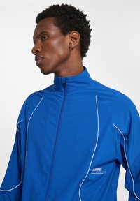 Blå zip-up jacka av strukturerad tyg, med vita accentlinjer och hög krage, samt en logotyp på vänster sida.