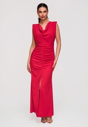 Femme portant une robe rouge sans manches froncée avec une fente sur le devant et un décolleté drapé, debout devant un fond clair uni.