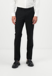 Pantalons noirs ajustés avec une texture lisse, coupe droite, et passants de ceinture, associés à des mocassins noirs et une chemise blanche à boutons.