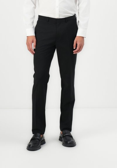Pantalons noirs ajustés avec une texture lisse, coupe droite, et passants de ceinture, associés à des mocassins noirs et une chemise blanche à boutons.