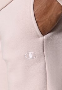 Sweat-shirt rose clair en tissu doux et texturé, avec un logo brodé blanc sur le côté gauche près de la poche.