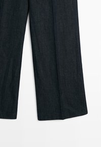 Tmavě modré džínové široké kalhoty visící, zobrazující celou délku obou nohavic na světlém pozadí.