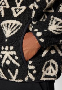 Jachetă din fleece neagră cu modele abstracte albe. Dispune de un buzunar lateral și o țesătură texturată. Încadrată cu țesătură neagră la margini.