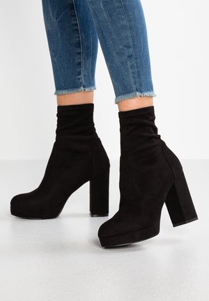 Czarne zamszowe botki za kostkę z wysokim klockowym obcasem, luźnym krojem cholewki i zaokrąglonym noskiem, zestawione z skróconymi niebieskimi dżinsami.