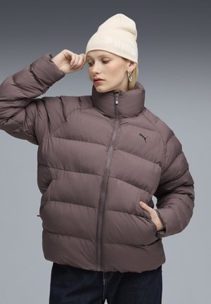 Vrouw past een crèmewitte Puma-beanie aan terwijl ze een bruine pufferjack en donkere jeans draagt, poserend tegen een grijs gradientachtergrond.
