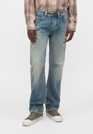 Persona que lleva vaqueros azules desgastados y rotos, camisa marrón de cuadros y zapatillas grises, de pie frente a un fondo blanco liso.