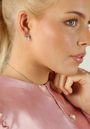 Silverörhängen i en dubbelögladesign med klara accenter, bärs av en person i en rosa satinblus och ett pärlhalsband.