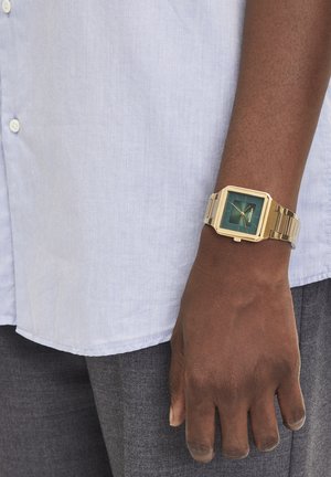 Goldfarbene rechteckige Uhr mit grünem Ziffernblatt, Metallband und kantigem Design. Am Handgelenk getragen, in Kontrast zu einem hellblauen Hemd.
