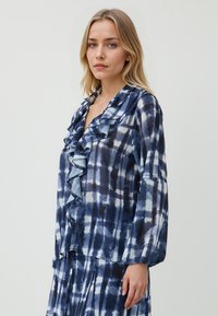 Blusa blu navy con placket arricciato, caratterizzata da un motivo tie-dye in tonalità di blu più chiare. Maniche lunghe e tessuto leggero.