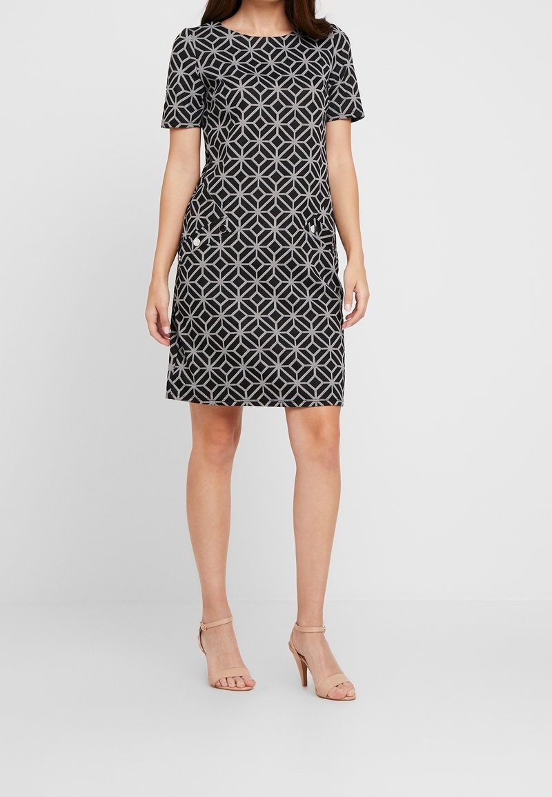 Femme portant une robe noire mi-longue avec un motif géométrique gris, manches courtes, poches, et sandales à talons hauts beiges, debout sur un fond uni.