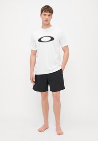 Weißes T-Shirt mit kurzen Ärmeln und einem schwarzen ovalen Logo, kombiniert mit schwarzen strukturierten Sportshorts mit Seitentaschen. Das Modell steht barfuß.