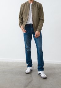 Mężczyzna stojący w pomieszczeniu, ubrany w oliwkową kurtkę, białą koszulkę, niebieskie dżinsy i białe trampki w czarne paski na betonowej podłodze.