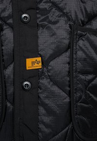 Black quilted jacket with a textured finish, featuring two front pockets, a gray button closure, and an orange brand tag.