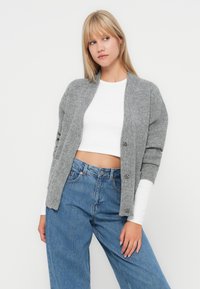 Junge Frau in einem grauen Strickcardigan über einem weißen verkürzten Top und hochgeschnittenen blauen Jeans, die vor einem weißen Hintergrund steht.