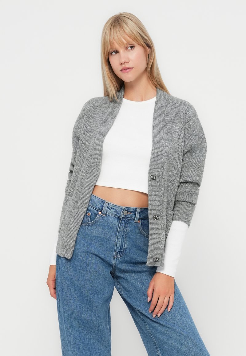 Giovane donna che indossa un cardigan grigio lavorato a maglia sopra un top bianco corto e jeans blu a vita alta, in piedi davanti a uno sfondo bianco.