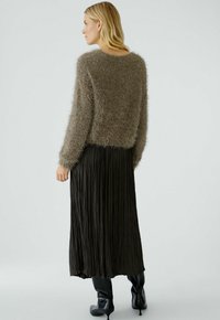 Un pull en laine marron et duveteux associé à une jupe maxi plissée noire et des bottes noires haute jusqu'aux genoux, vu de dos.