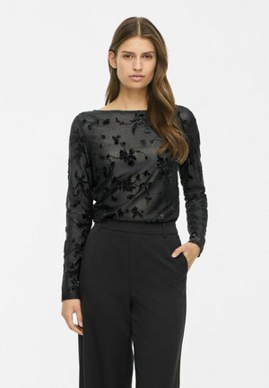 Un mannequin porte un chemisier noir transparent à manches longues orné de motifs floraux et un pantalon noir, et se tient devant un fond uni.