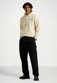 Kremna flis kapuca z žepom spredaj in črnim logotipom, kombinirana s črnimi cargo hlačami. Model nosi črne superge. Preprost dizajn.