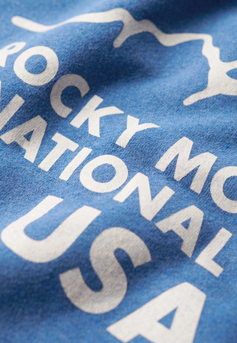 Camiseta de algodón azul con texto blanco que dice "PARQUE NACIONAL ROCKY MOUNTAIN EE. UU.", con una sutil textura y un gráfico de montañas encima del texto.
