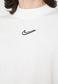 Camiseta blanca de manga larga con cuello alto, que presenta un logo bordado de Nike en negro en el pecho. Textura de tela suave.