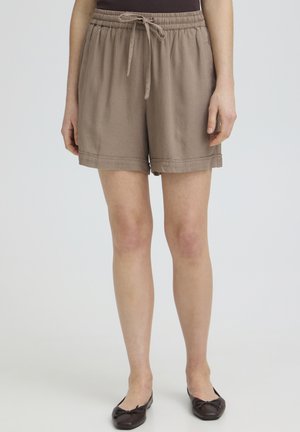 Løse taupe-shorts med elastisk taljebånd og snor, båret med mørke balletsko på en person, der står mod en lys baggrund.