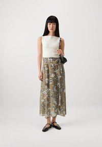 Ærmeløs, ribstrikket hvid top kombineret med et langt nederdel med blomstermønster i guld- og blå toner. Modellen holder en lille sort håndtaske.