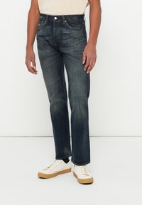 Jeans de mezclilla azul oscuro con pernera recta, ligero desgaste, diseño de cinco bolsillos y un cierre clásico de botón y cremallera. Llevados con zapatillas blancas.
