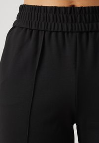 Närbild av svarta elastiska midjebandsbyxor med en slät tyg och subtil sömdetalj på framsidan.